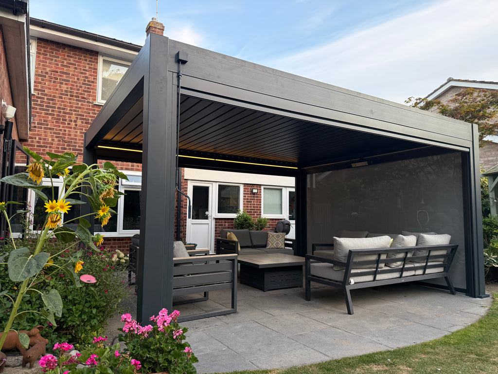A modern dark gray outdoor patio cabana with a slatted roof, featuring a built-in screen wall, a coffee table, and a cushioned bench. The cabana is set in a backyard with a brick house, green plants, and flowers in the foreground.
Details
