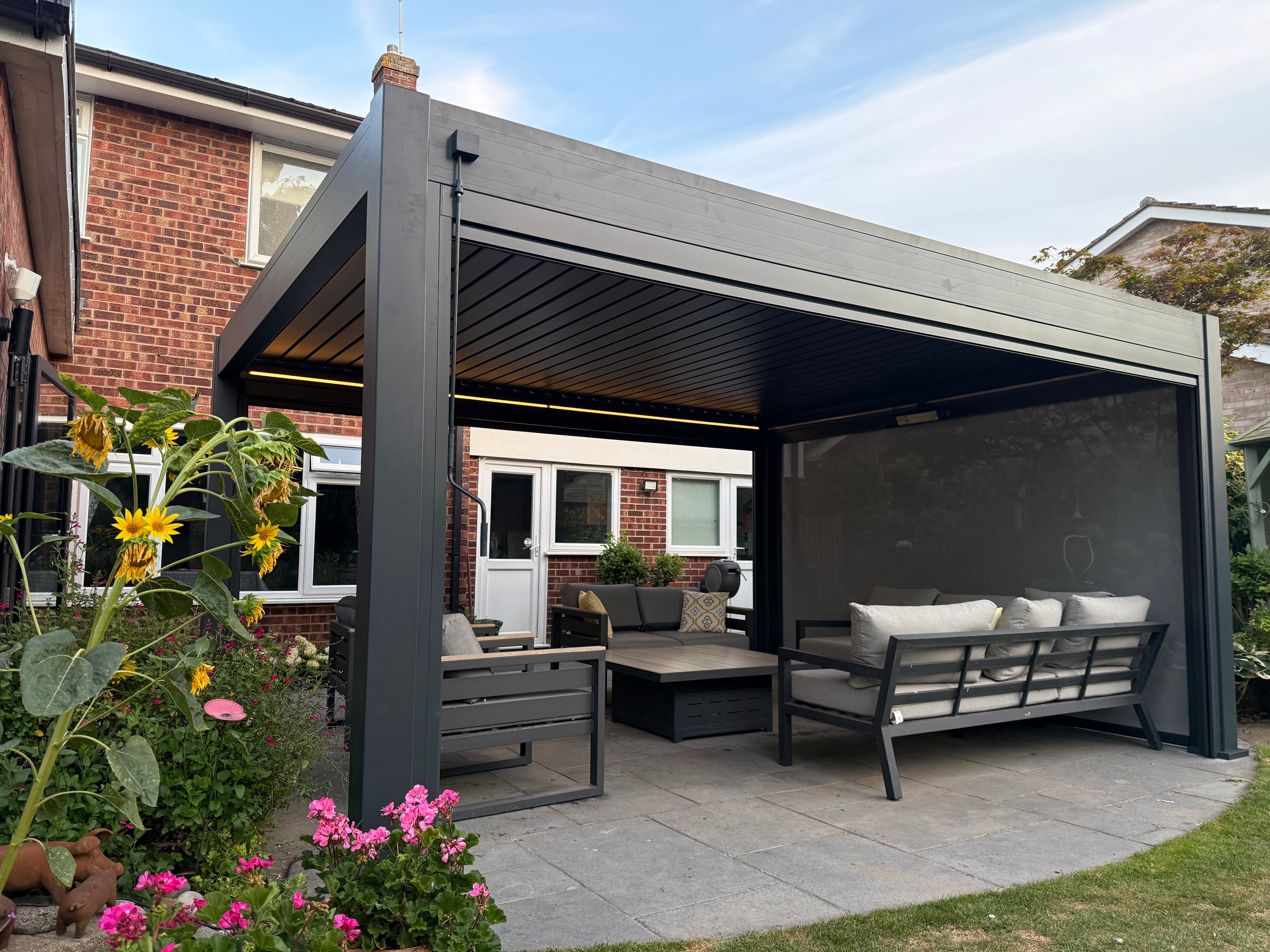 A modern dark gray outdoor patio cabana with a slatted roof, featuring a built-in screen wall, a coffee table, and a cushioned bench. The cabana is set in a backyard with a brick house, green plants, and flowers in the foreground.
Details